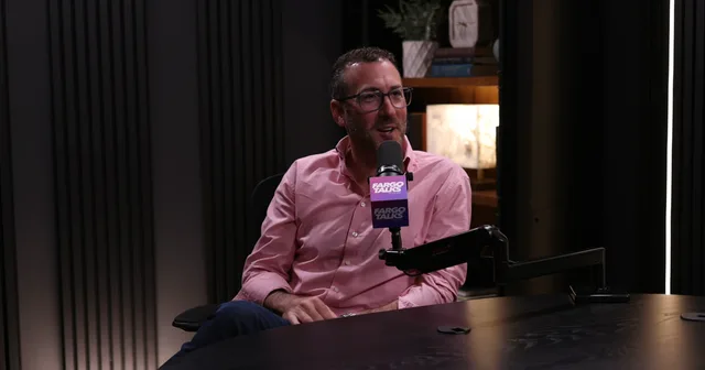 Judd Hoffman in a pink shirt speaking into a Fargo Talks microphone in a dark wood-panelled podcast studio, mid-conversation.